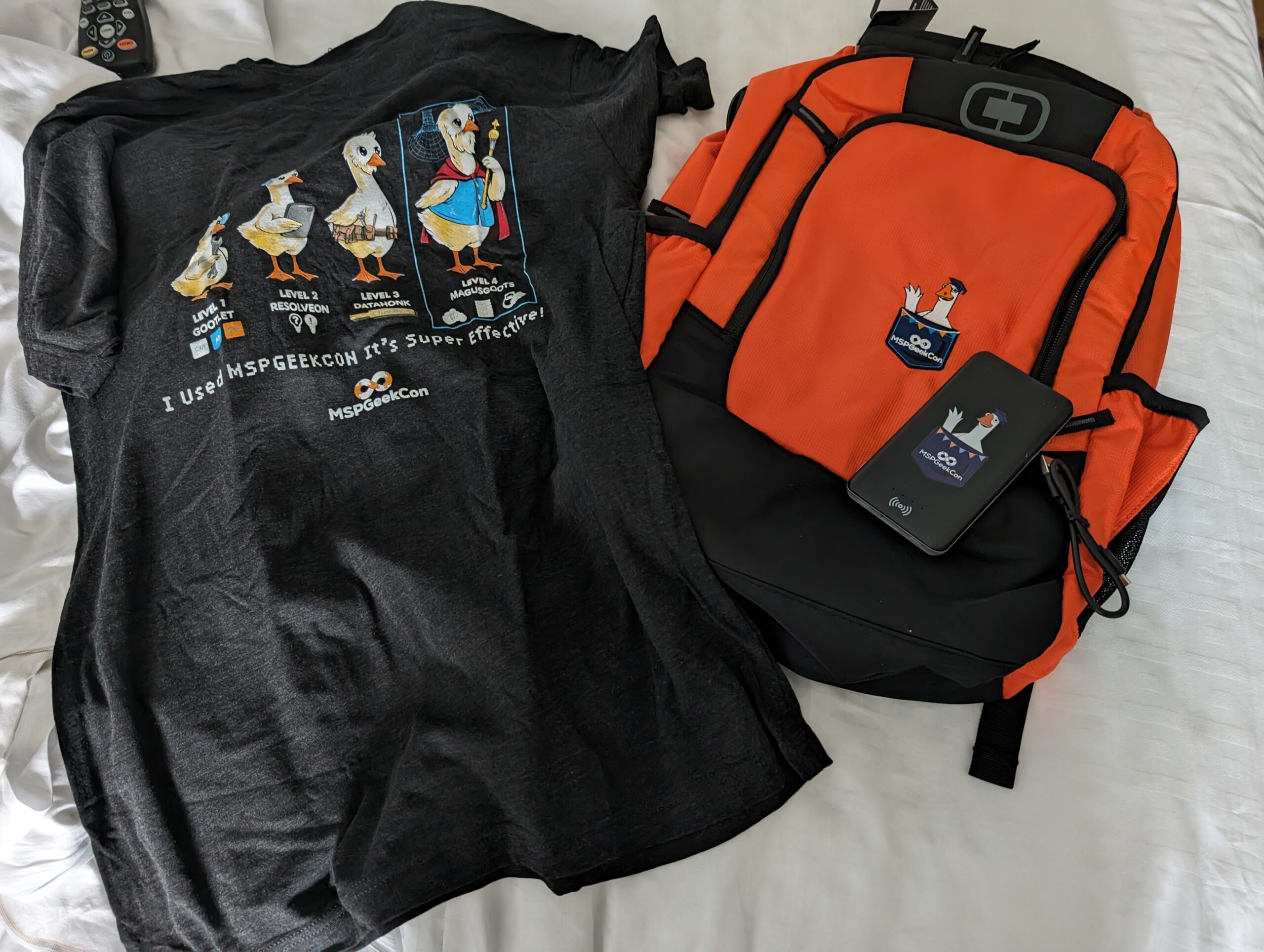 orange backpack, black tshirt, and a black power battery bank on a bed. Each item has a cartoon goose on it. The tshirt has four geese in varying stages of "technical evolution"